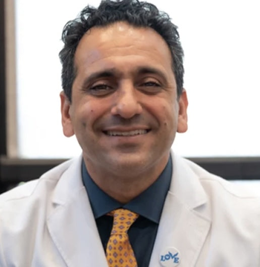 A man wearing a white lab coat, a dark shirt, and a patterned yellow tie is smiling at the camera.