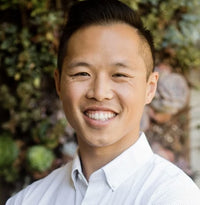 A person smiling, wearing a white collared shirt, standing in front of a blurred background with green and brown plants.