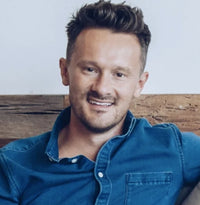 A man with short brown hair and a trimmed beard, wearing a blue button-up shirt, sits and smiles in front of a wooden wall.