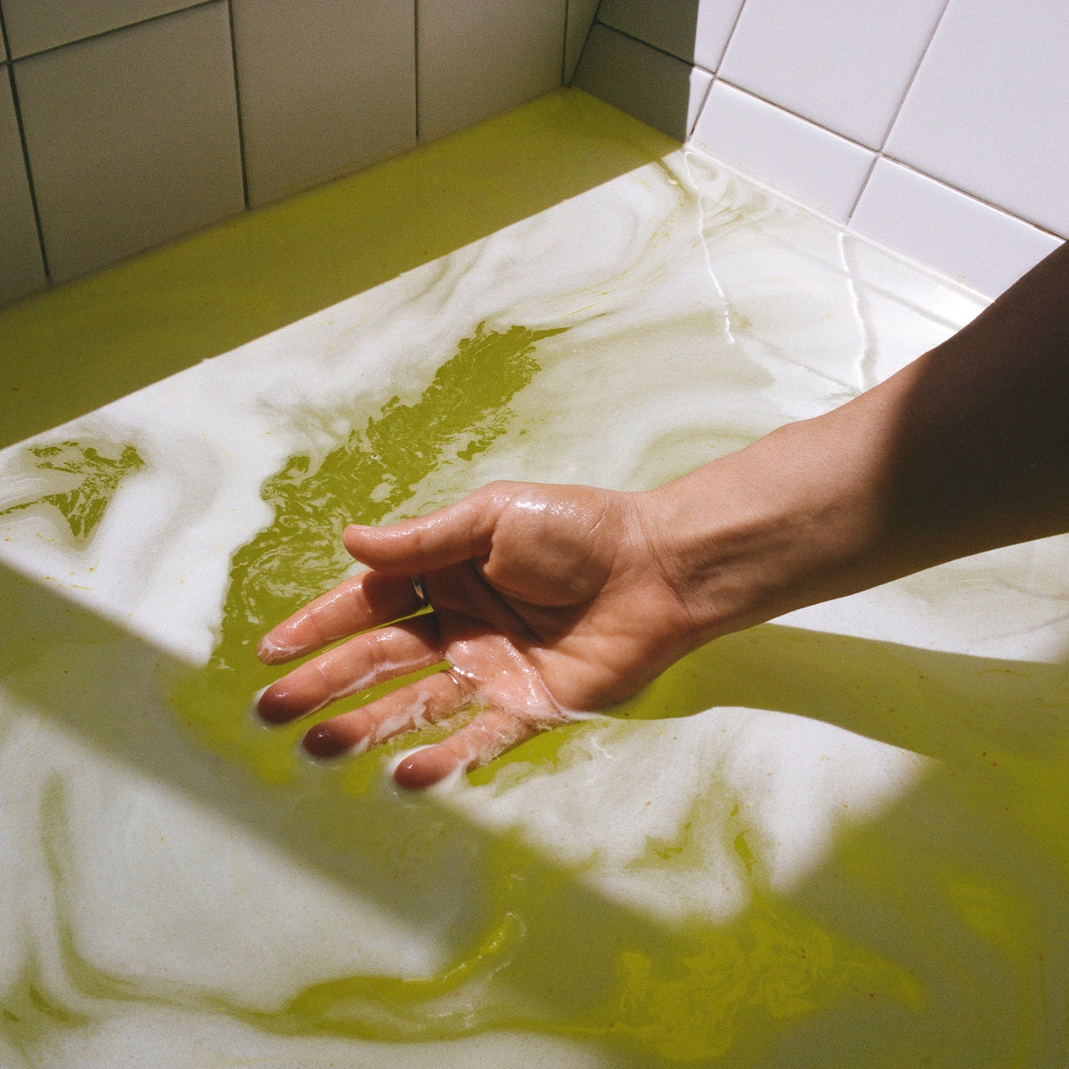 A hand touching a yellow-green colored water with THE FULLEST Exheal salt in a sunlit bathtub.