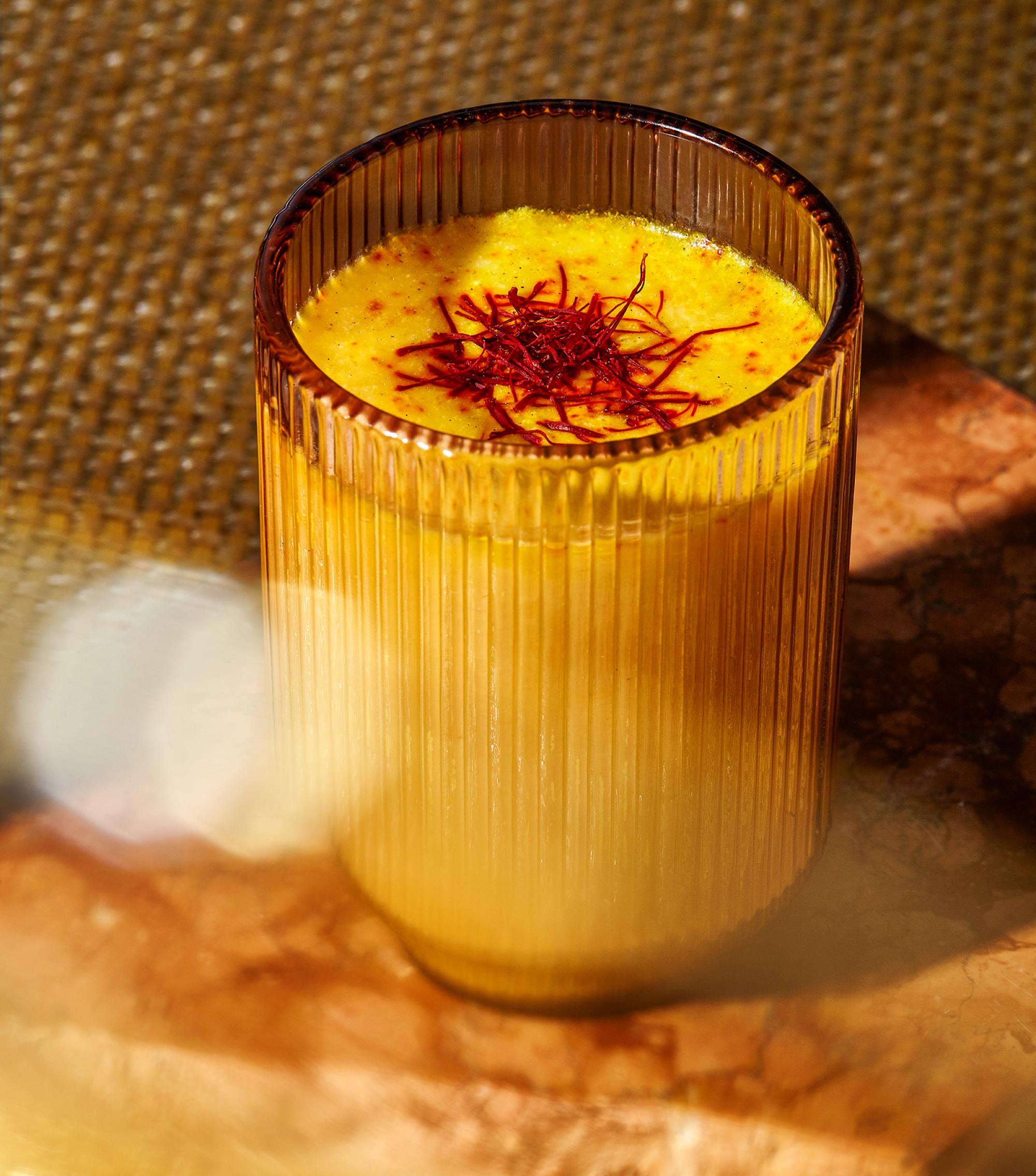 A glass of THE FULLEST Warm Feelings, a golden-hued caffeine-free latte garnished with red saffron strands, on a textured surface.