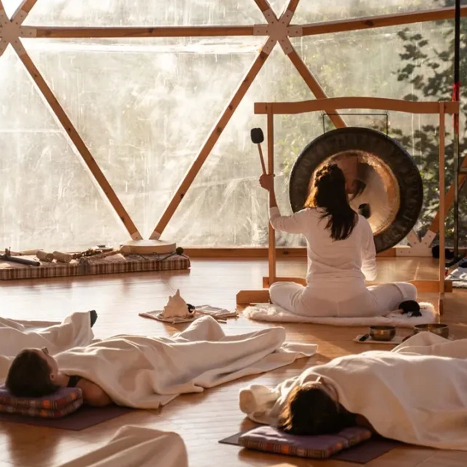 A Visionary Sound Bath session with participants lying down and an instructor playing a gong in a room with large windows and natural light, inspired by the serene vibes of Laguna Beach. Brand Name: THE FULLEST.