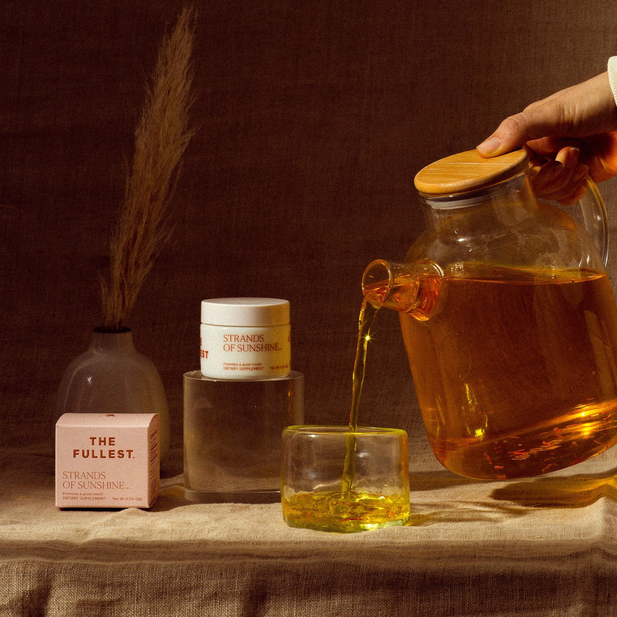 A model pouring a golden saffron tea from a glass tea pot into a small cup on a draped cloth surface, with decorative bottles and Strands of Sunshine threads nearby by THE FULLEST.