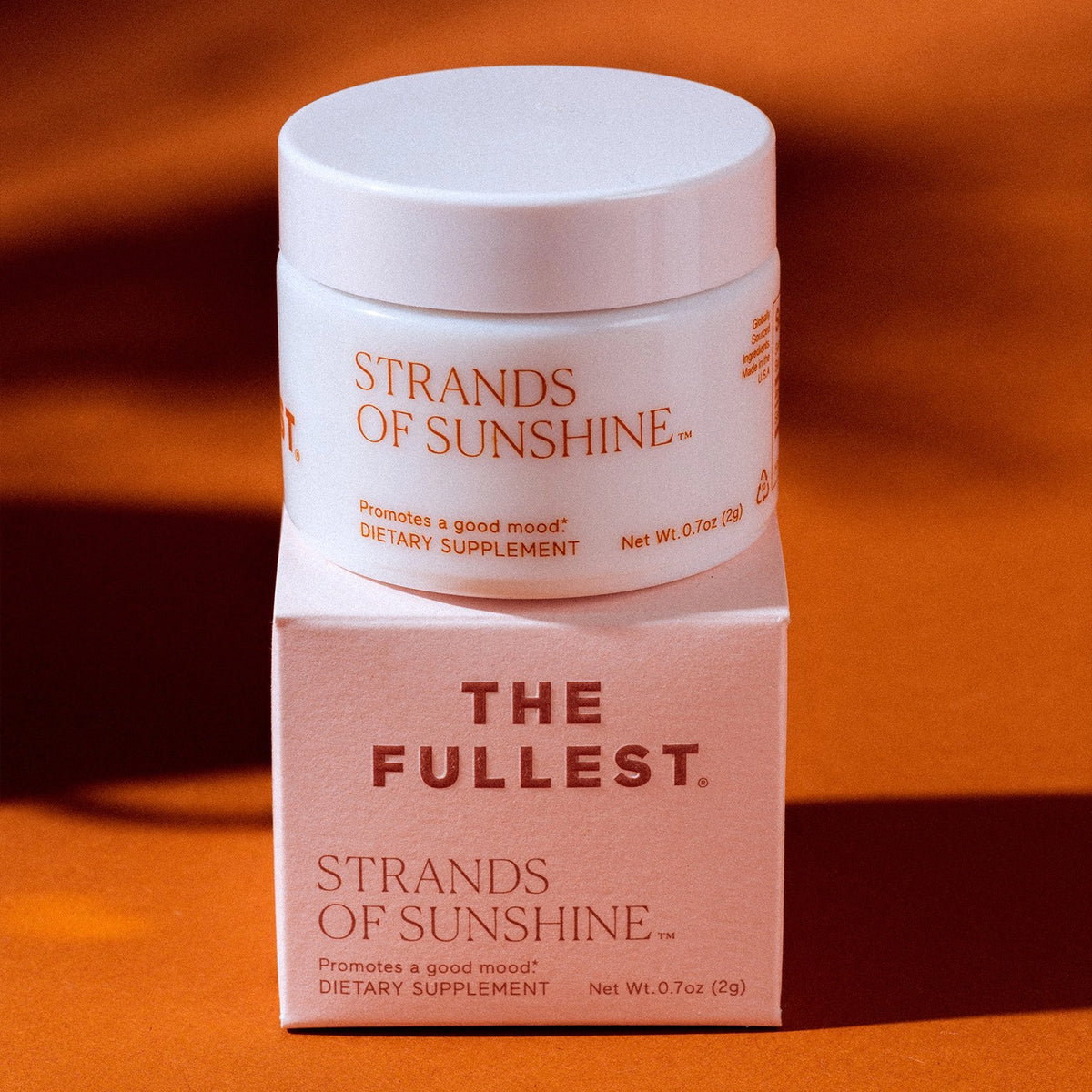 A bottle of &quot;Strands of Sunshine&quot; by THE FULLEST containing pure saffron flower threads, resting on top of its packaging box against an orange backdrop.