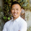 A man with short dark hair wearing a white button-up shirt smiles with arms crossed, standing in front of a leafy background.