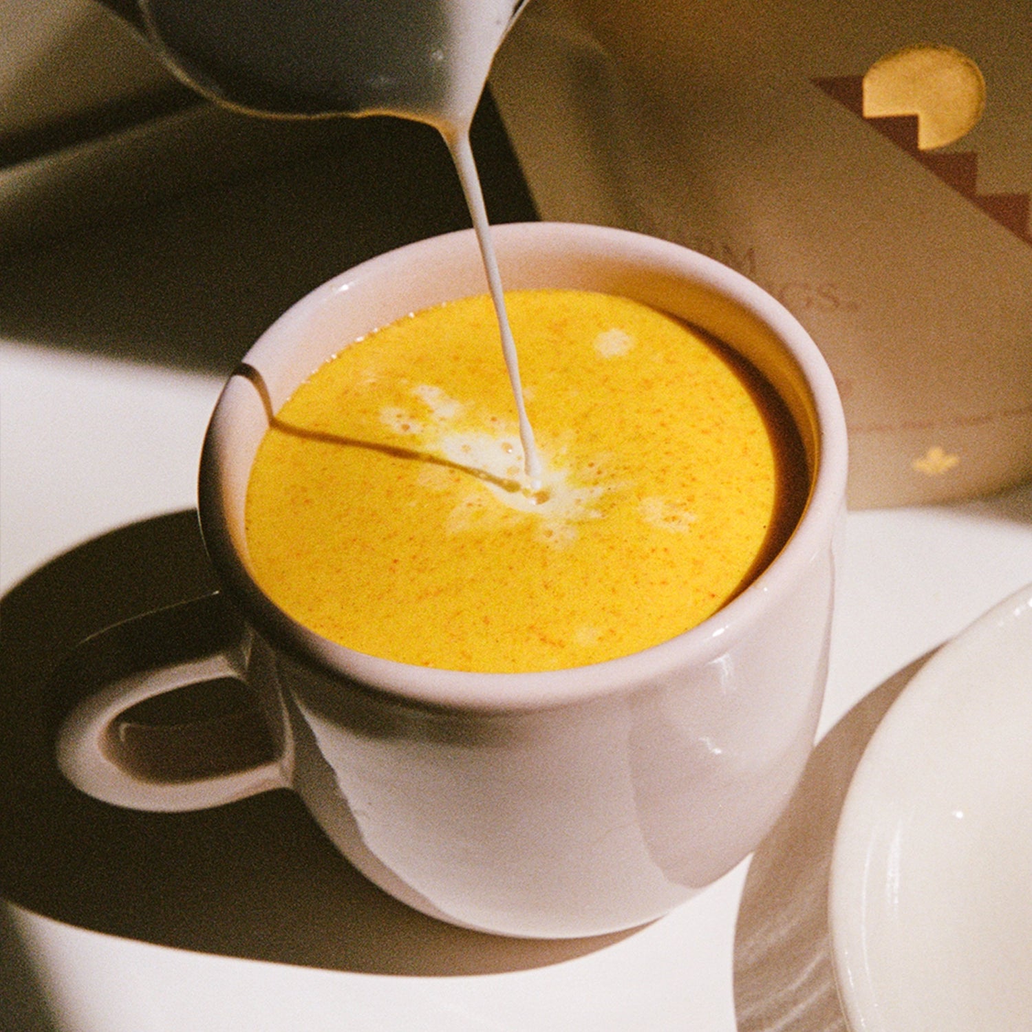 A white mug filled to the brim with a vibrant, saffron-colored frothy drink, as a stream of milk is being poured in, creating a delicate pattern on the surface. The mug sits next to a saucer.
