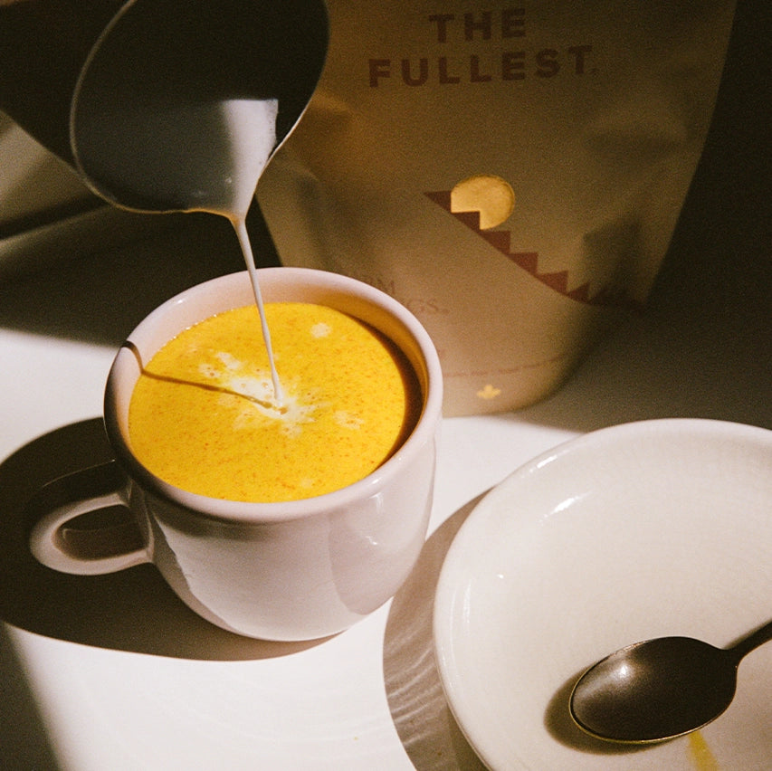 A white mug filled to the brim with a vibrant, saffron-colored frothy drink, as a stream of milk is being poured in, creating a delicate pattern on the surface. The mug sits next to a saucer.