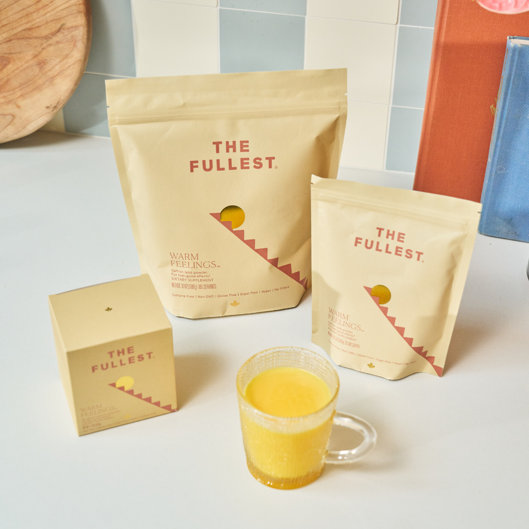 Three beige The Fullest saffron latte bags and a 10-sachet box next to a clear glass mug of golden saffron latte on a white countertop.