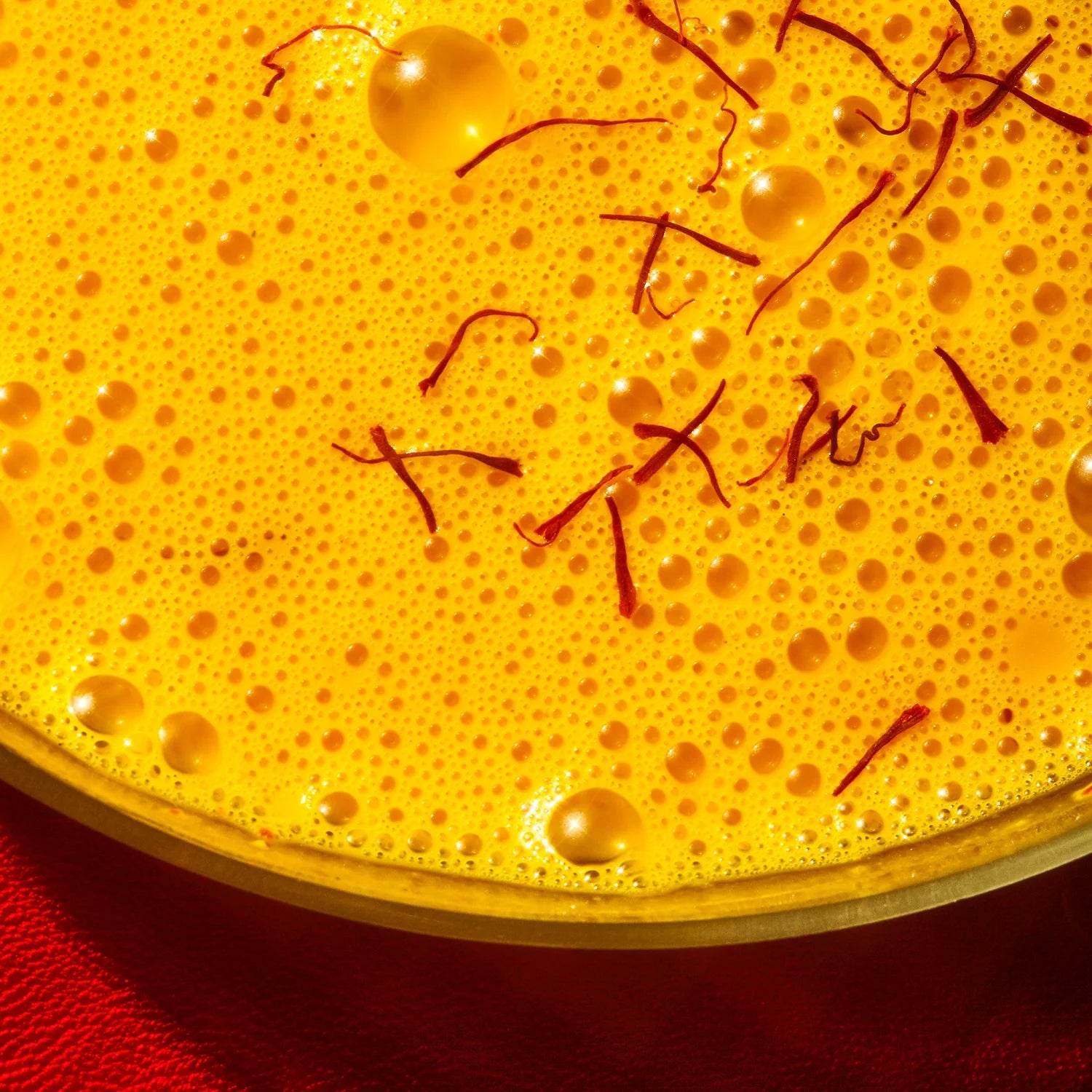 A close-up of THE FULLEST's Saffron Bundle latte, with a yellow frothy surface and saffron strands on top, served in a glass against a red background.